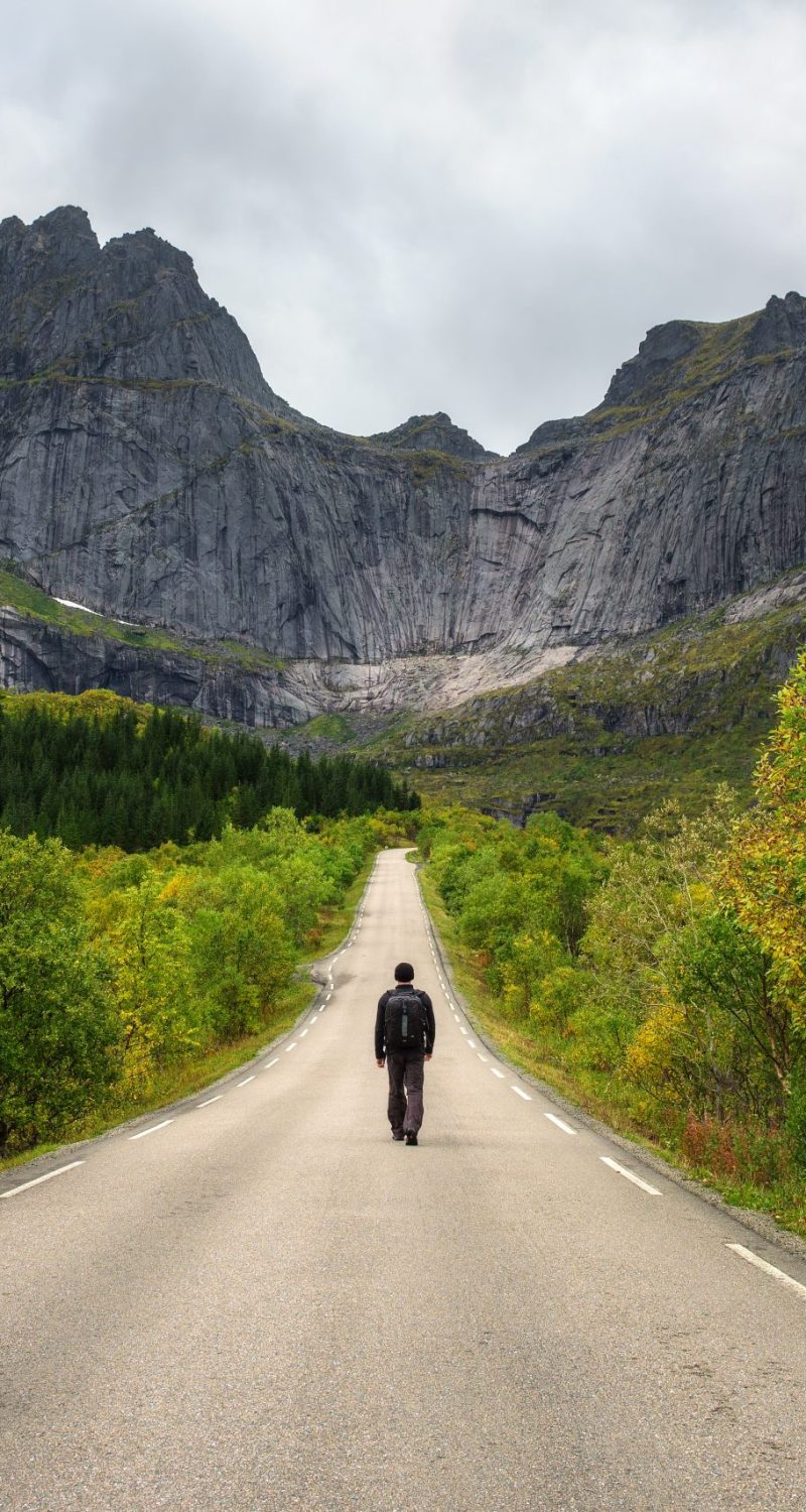 Hiker carrying a backpack walks on a scenic road on Lofoten islands in Norway. Concept for loneliness and relaxation.